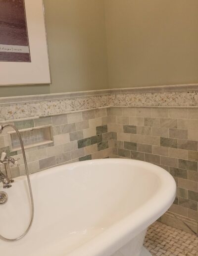 A white freestanding bathtub with silver fixtures, surrounded by beige and green tiled walls in a bathroom.