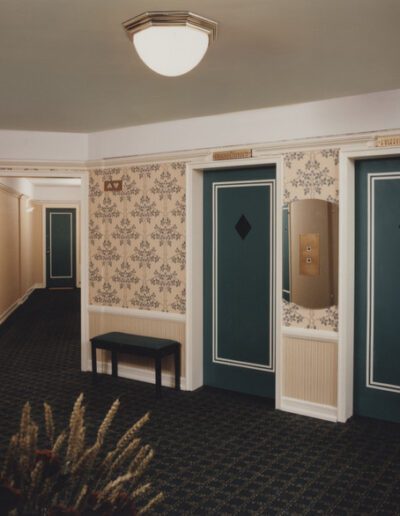 A traditional hallway with decorative wallpaper, two teal doors, benches, and a carpeted floor.