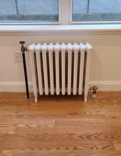A traditional white radiator heater installed below a window in a room with hardwood flooring.