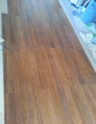 A wooden floor with a warm brown finish, showing signs of use, with some clutter visible in the background.